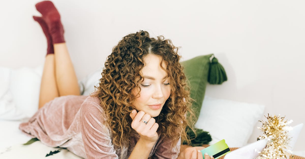 A woman with curly hair shops online on a laptop, lying on a bed decorated with tinsel and string lights.