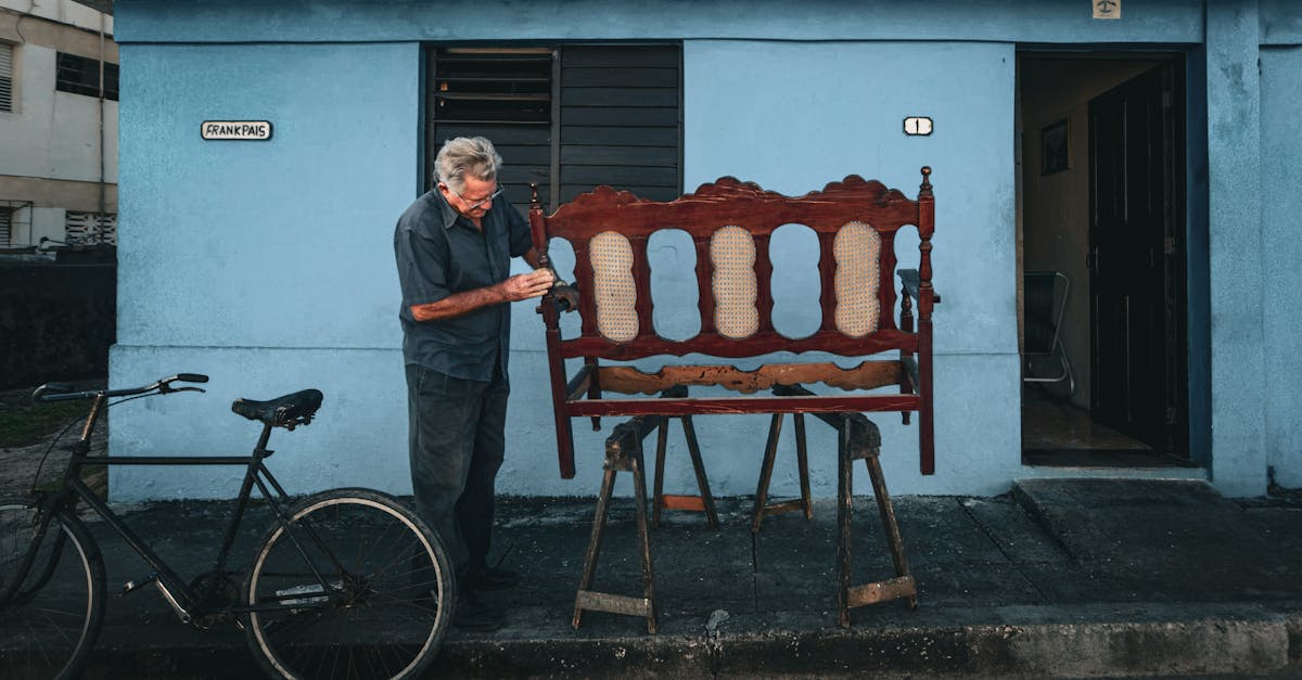 Upcycling furniture - An elderly man works on restoring a vintage wooden bench outside a blue house.