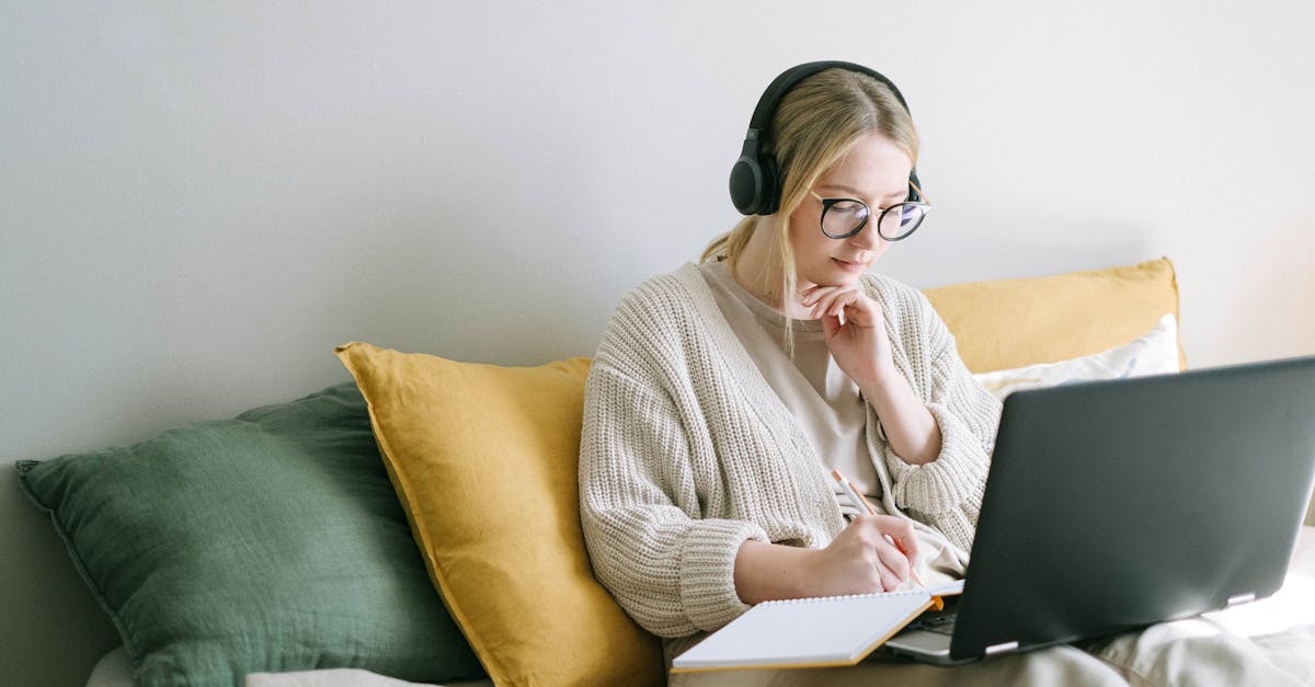 Income generation through blogging - A focused woman in glasses and headphones works on a laptop from a cozy bed.